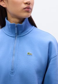 Μπλε φούτερ με φερμουάρ, κατασκευασμένο από υφασμα με υφή, με ψηλό γιακά και μικρό πράσινο λογότυπο κροκοδείλου στο στήθος.