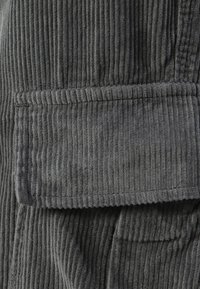 Šedá kordurová látka s vertikálním žebrováním, která obsahuje klopovou kapsu s prošívaným detailem podél okraje. Měkká textura, jednotný vzor.