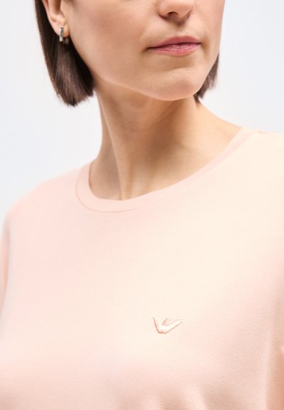 Γυναίκα που φοράει ένα ανοιχτό ροζ μπλουζάκι με στρογγυλή λαιμόκοψη, με ένα μικρό κεντητό λογότυπο στο στήθος και ασημένια κρίκους σκουλαρίκια.