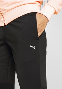Main dans la poche d'un pantalon noir Puma porté avec une veste zippée couleur pêche, montrant le logo Puma sur la cuisse.