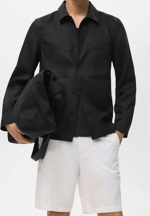 Osoba w czarnej koszuli na guziki i białych szortach, trzymająca czarną złożoną torbę w jednej ręce, druga ręka w kieszeni na białym tle.