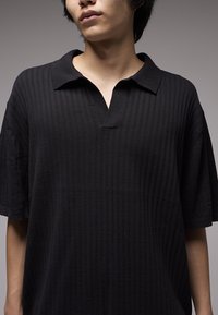 Μαύρο πλεκτό πόλο πουκάμισο με ανάγλυφη υφή, κοντά μανίκια και σχεδιασμό γιακά με σχίσιμο. Απλός και κλασικός στιλ χωρίς επιπλέον διακοσμητικά στοιχεία.