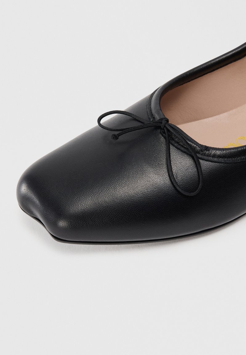 Ballerines en cuir noir avec un bout carré, ornées d'un fin nœud en dentelle sur le dessus, texture lisse, et semelle intérieure beige.