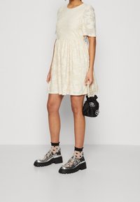 Cremefarbenes Kleid mit einem strukturierten Blumendesign, kurzen Ärmeln, gerafftem Taillenbund, kombiniert mit silbernen Plateauschuhen und einer schwarzen Tasche.