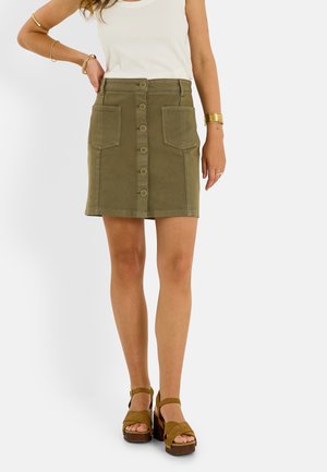 Mujer con top blanco sin mangas, minifalda verde oliva con botones delanteros y bolsillos, y sandalias de plataforma verde oliva con tiras en el tobillo.