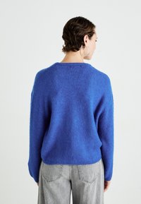 Woman with short wavy hair wearing large hoop earrings, a blue fuzzy sweater, and light gray jeans, facing away against a plain background.