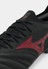 Μαύρο ποδοσφαιρικό παπούτσι με μαύρες κορδόνες και ένα μεγάλο κόκκινο στυλιζαρισμένο λογότυπο στο πλάι, εμφανίζεται σε κοντινή λήψη σε λευκό φόντο.