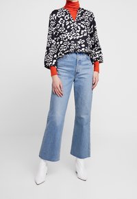 Czarna i biała bluzka w wzory z szerokimi rękawami na czerwonym golfie, połączona z jasnoniebieskimi spodniami z wysokim stanem w stylu dzwonów i białymi botkami do kostek.