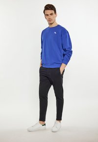Felpa blu reale con maniche lunghe, scollo rotondo e piccolo logo; abbinata a pantaloni neri slim-fit e sneakers bianche con dettagli verdi.