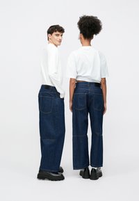 Τζιν παντελόνια με φ�αρδιά μπατζάκια σε σκούρο τζιν ύφασμα, με διακριτές τσέπες και ραφές σε αντίθετο χρώμα. Τα μοντέλα φοράνε λευκά τοπ και χοντρά μαύρα παπούτσια, απεικονίζονται από πίσω.