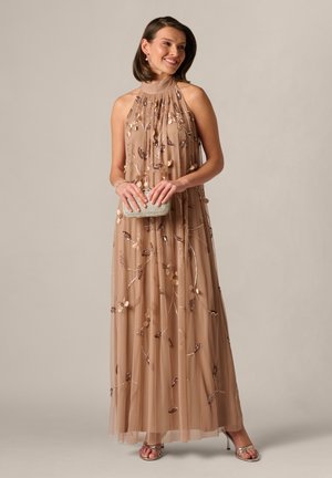 Kobieta uśmiechająca się, ubrana w długą beżową suknię na szyję z kwiatowymi cekinami, trzymająca małą błyszczącą kopertówkę, w srebrnych szpilkach i z krótkimi włosami.