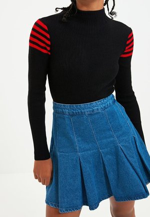 Personne portant un pull noir côtelé à manches longues avec des rayures rouges sur les épaules et une jupe plissée en jean bleu.