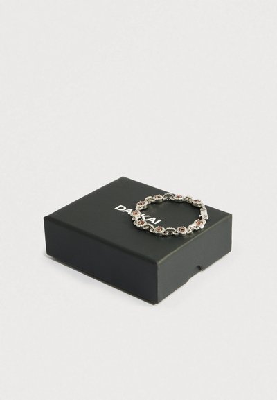 Ezüst karkötő váltakozó barna és tiszta drágakövekkel, fekete dobozon, fehér "DAKAI" felirattal, egyszerű világos háttér előtt.