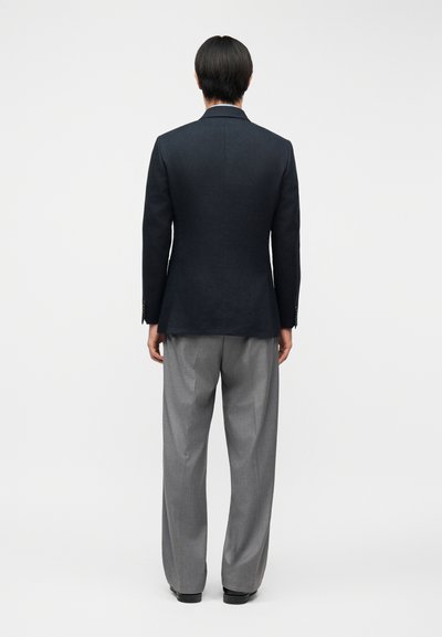 Άνδρας με την πλάτη γυρισμένη φοράει μαύρο σακάκι, γκρι πιετέ παντελόνια και μαύρα παπούτσια, στέκεται μπροστά σε ένα απλό ανοιχτό γκρι φόντο.