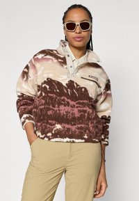 Fleece pulóver v béžovej, hnedej a ružovej vzoroch, s vysokým golierom a zipsovou vreckom. Nosený s svetlohnedými nohavicami a slnečnými okuliarmi.