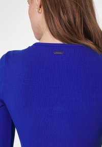 Femme aux longs cheveux bruns portant un haut côtelé bleu royal avec un logo Guess en métal dans le haut du dos et une boucle d'oreille en or.