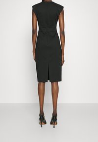 Mustissa polvipituisessa mekossa on jämäkät olkapäät, takareikä ja muotoiltu siluetti. Se on valmistettu sileästä kankaasta ja yhdistetty mustiin teräväkärkisiin korkokenkiin.