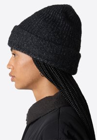 Berretto lavorato a maglia nero con una texture a coste, dotato di un risvolto. La vista laterale mostra capelli neri intrecciati e una giacca nera con colletto sottostante.