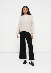 Cardigan de punto beige con cierre de botones, mangas anchas, usado sobre una blusa rosa clara y combinado con pantalones de pierna ancha negros y mocasines negros.