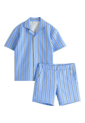 Ung dreng smilende, iført blå og hvide vertikale stribede kortærmede skjorte og matchende shorts, blå kasket, sokker og marineblå sneakers.