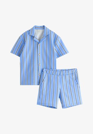 Ung dreng smilende, iført blå og hvide vertikale stribede kortærmede skjorte og matchende shorts, blå kasket, sokker og marineblå sneakers.