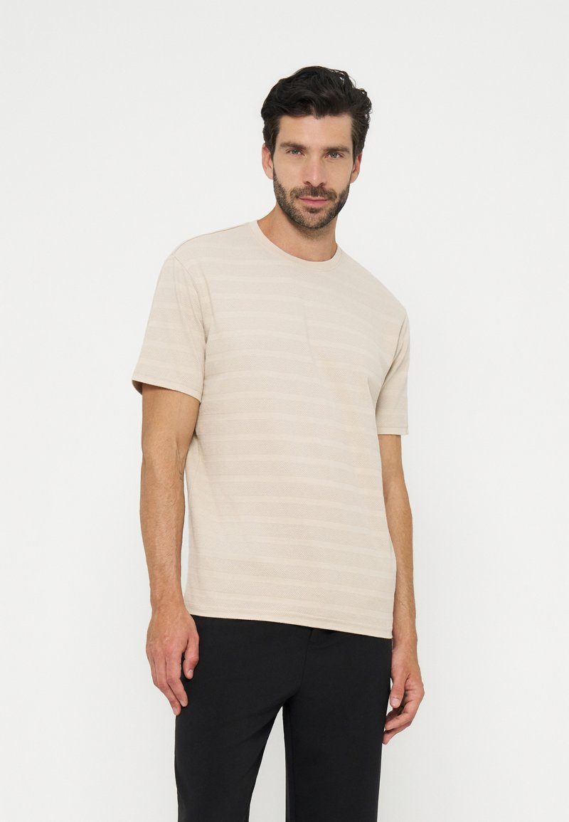 Homme aux cheveux foncés et à la barbe, portant une chemise beige texturée à manches courtes et un pantalon noir, debout devant un fond blanc.