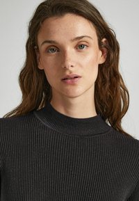 Maglione nero a coste con collo alto. Il materiale presenta una leggera lucentezza e la texture appare morbida ed elastica.