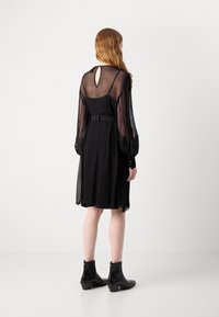 Robe noire avec des manches longues transparentes, taille ceinturée et détail de dos en ouverture. Associée à des bottines noires à talons hauts. Design simple, look élégant.