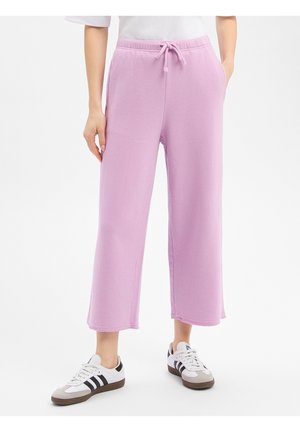 Person trägt helllila, weit geschnittene, cropped Jogginghosen mit seitlichen Taschen und weiße Turnschuhe mit schwarzen Streifen.