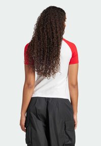 T-shirt blanc avec des manches raglan rouges, offrant une texture lisse. Le dos présente des cheveux bouclés dévalant sur le t-shirt.