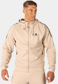 Beige zip-up hoodie van textiel met een gestructureerde stof, verstelbare trekkoordkap, zijzakken en zwarte logodetails op de borst en mouwen.