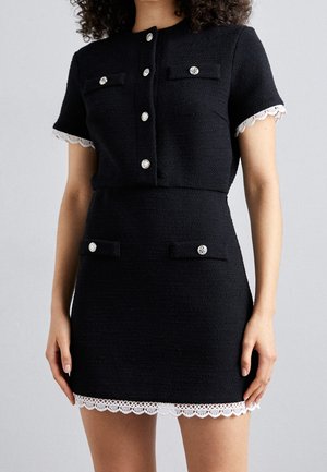 Woman wearing a fitted black short-sleeve dress with silver buttons, flap pockets, and white lace trim on sleeves and hem.