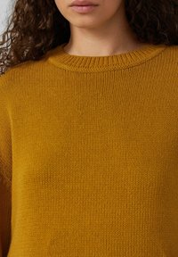 Musztardowy żółty sweter dzianinowy o teksturowanym, prążkowanym wzorze i klasycznym okrągłym dekolcie. Materiał wydaje się miękki i ciepły.