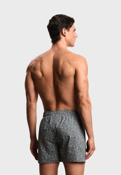 Άνδρας εμφανίζεται από πίσω, φοράει γκρι μπόμεξερ με σχέδια, στέκεται μπροστά σε απλό ανοιχτό γκρίζο φόντο.