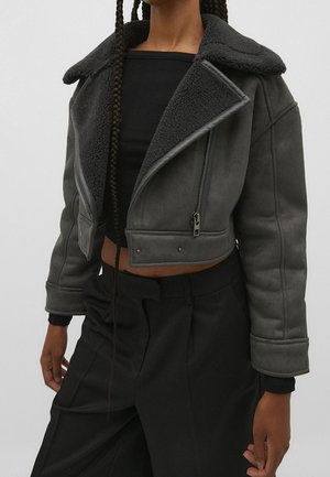 Veste courte noire en shearling avec col large et fermeture éclair argentée portée sur un haut court noir et un pantalon noir.