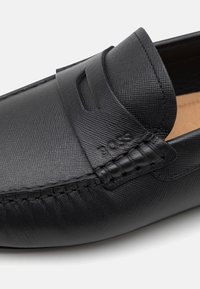 Mocassins en cuir noir présentant une texture, une couture de style mocassin, et une sangle élégante sur le devant avec un détail de logo.