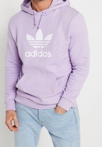 Světle fialová mikina s kapucí z měkké látky, na hrudi velké bílé logo Adidas a tři pruhy; obsahuje přední klokaní kapsu.