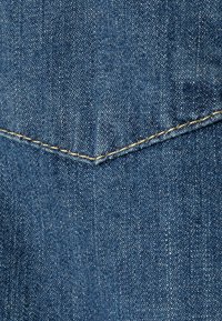 Denimtyg i mellanblått med en slät textur. Har en distinkt triangulär fickdesign och förstärkt sömnad i guld.