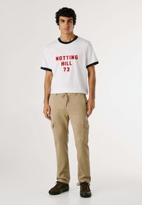 Vit t-shirt med röd text "NOTTING HILL 73", marinblå kant; beige cargobyxor med fickor; mörkbruna skor. Enkelt, avslappnat design.