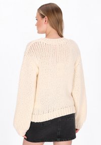 Cremfarbener Strickpullover mit lockerer Passform, grober Textur, raglanärmeln und gerippten Bündchen. Kombiniert mit einem schwarzen Denim-Minirock.