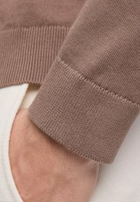 Brauner Strickpullover-Ärmelsaum mit geripptem Bündchen, das eine weiße Innenschicht zeigt. Fokus auf Textur und Farbkontrast.