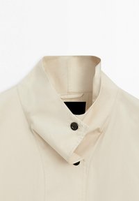 Beige jas met hoge kraag en zwarte knopen, voorzien van een opgevouwen kraag en zichtbare stiksels op een witte achtergrond.