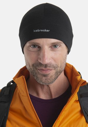 Schwarze Mütze mit nahtlosem Design, weicher Textur und "Icebreaker"-Branding, getragen von einer Person in einer orangefarbenen Jacke mit Reißverschluss und dunklem Hemd.