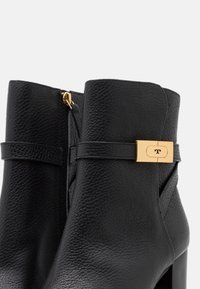 Botas de tobillo de cuero negro con un acabado texturizado, que presentan un accesorio de hardware dorado y un cierre con cremallera lateral. Diseño de tacón grueso.