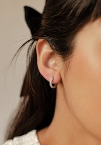 Créole en argent avec une forme courbée et une surface scintillante, portée sur une oreille visible, avec des cheveux foncés partiellement couvrant l'arrière-plan.