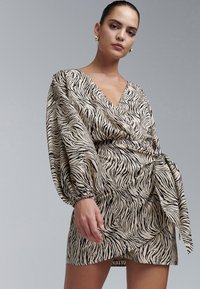 Sieviete valkā bēša un melnas zebras raksta wrap kleitu ar apjomīgām piedurknēm un zelta riņķveida auskariem, uz vienkārša pelēka fona.