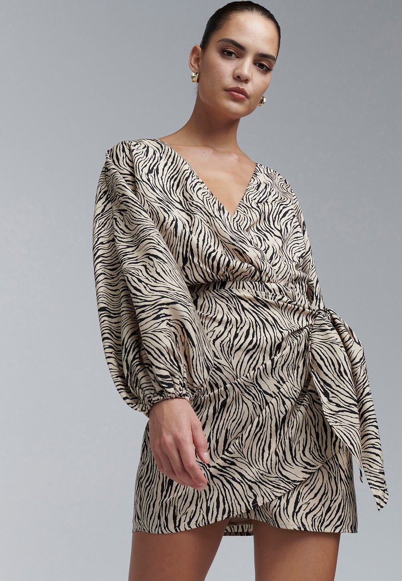 Sieviete valkā bēša un melnas zebras raksta wrap kleitu ar apjomīgām piedurknēm un zelta riņķveida auskariem, uz vienkārša pelēka fona.