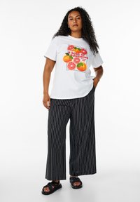 T-shirt bianca con grafica di frutti colorati e testo, abbinata a pantaloni a gamba larga a righe verticali neri e sandali neri. Capelli ricci.