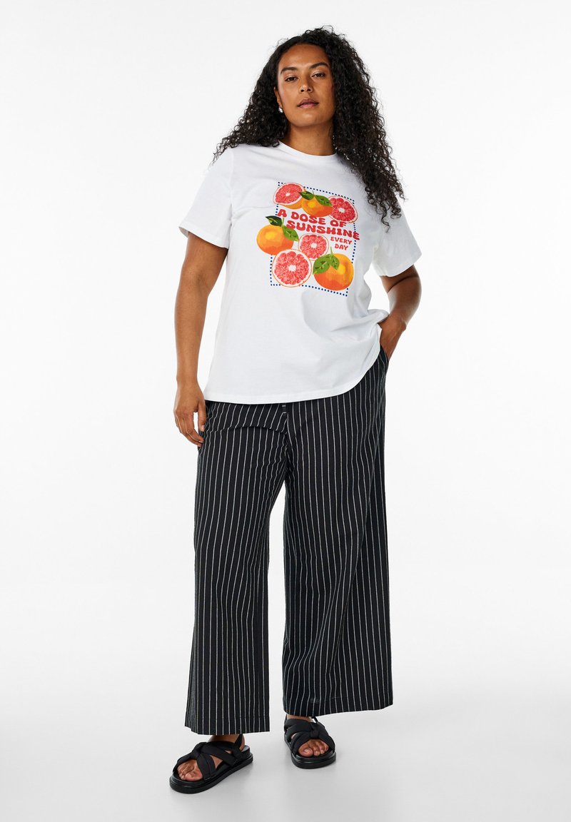 Vit t-shirt med färggrafik av frukter och text, kombinerad med svarta vertikala randiga vida byxor och svarta sandaler. Lockigt hår.
