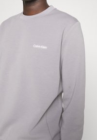 Felpa grigia in cotone con collo a giro, caratterizzata da un piccolo logo bianco "Calvin Klein" sul lato sinistro del petto. Texture liscia, design classico.
